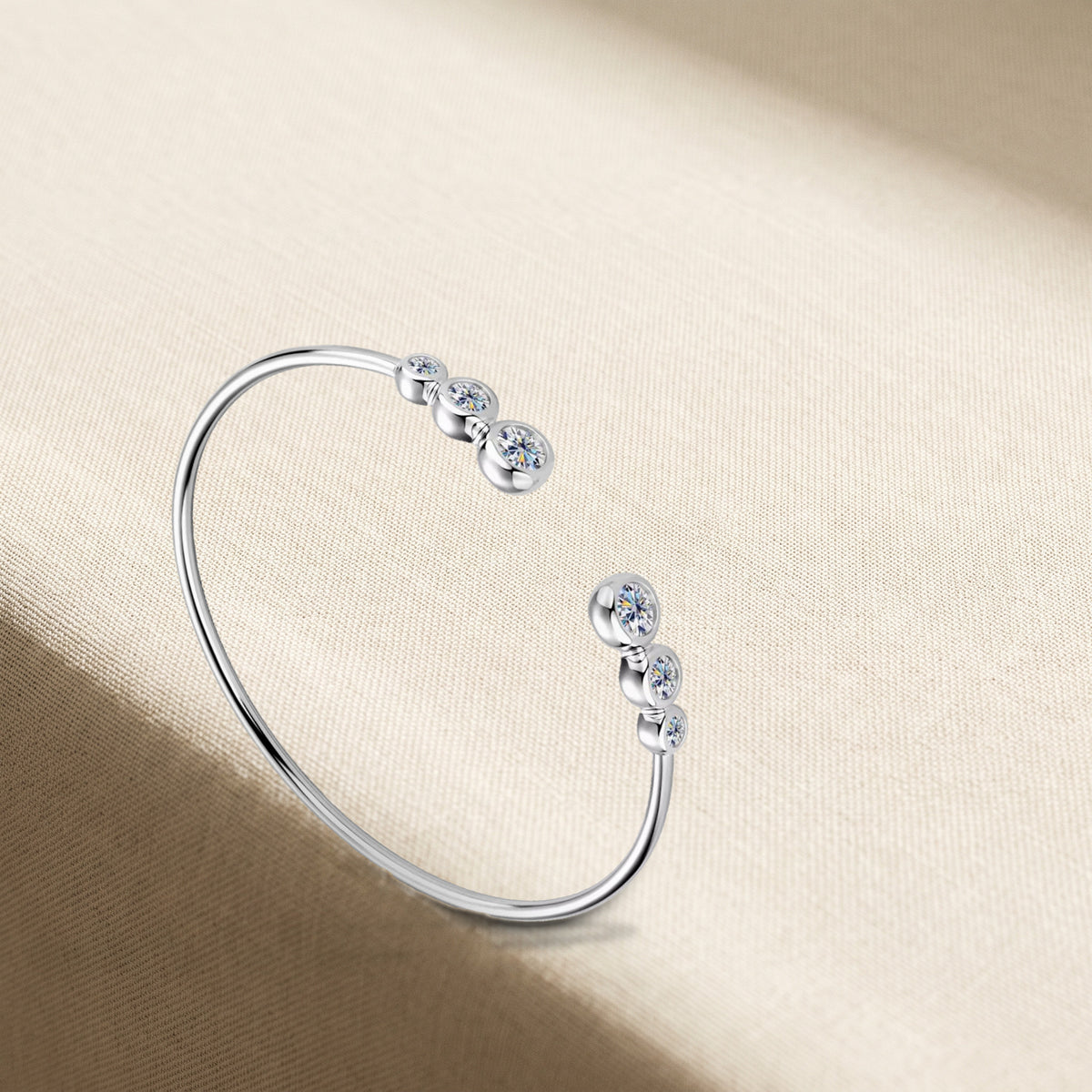 The Mossante Bezel Cuff on warm cream linen in soft window light: an open cuff of smooth round silver wire, each end finished with three round moissanites in rounded closed bezels, largest at the tip, the two ends facing each other across the opening.