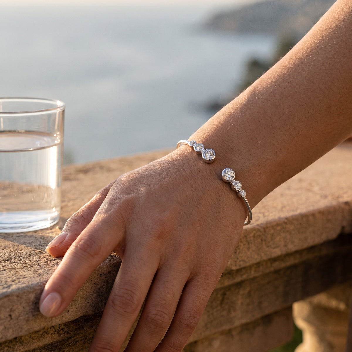 A woman's hand resting on a warm stone balcony ledge in late sun beside a glass of water, the sea behind, the silver Bezel Cuff's six stones on top of her wrist.