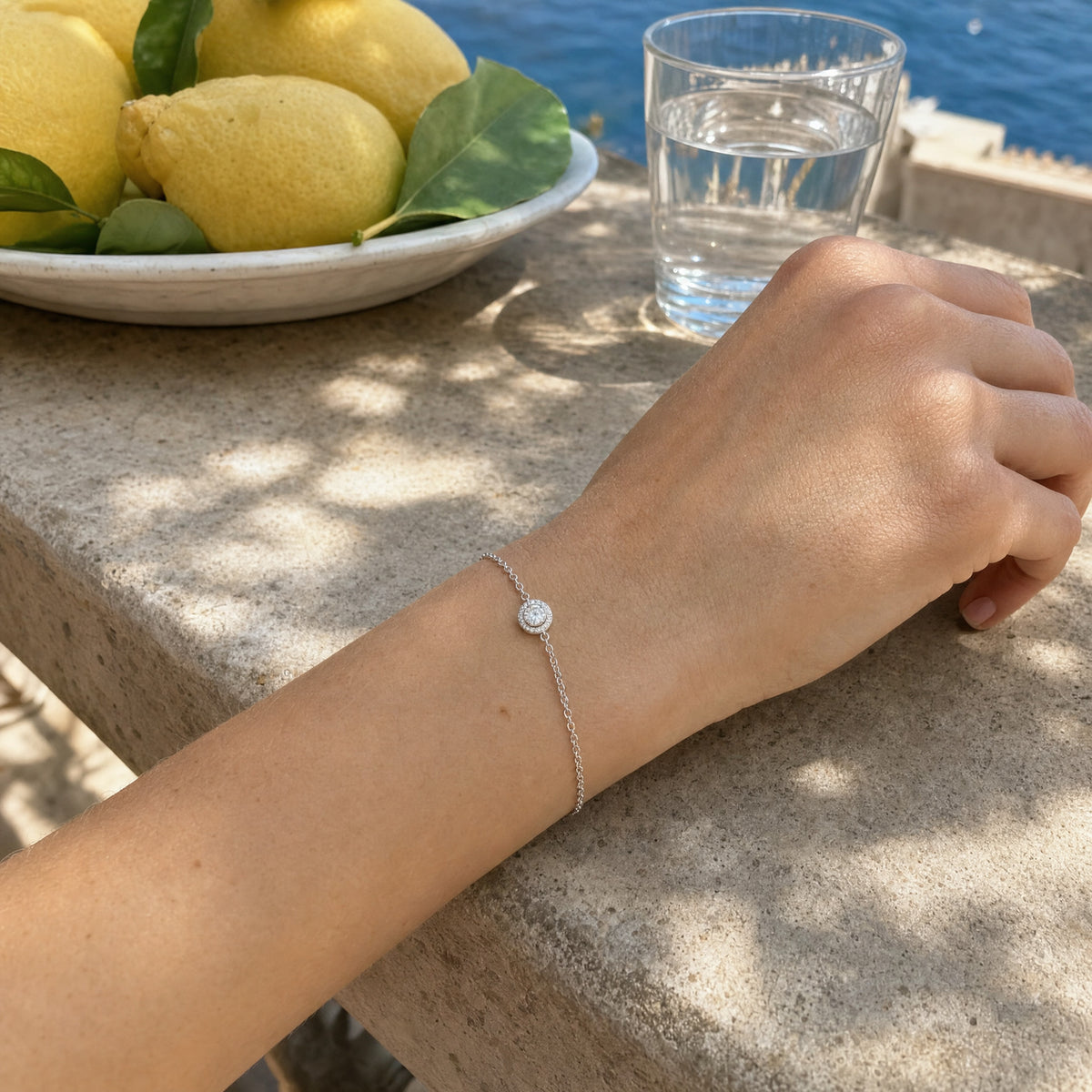 Her hand resting on a warm stone balustrade beside a bowl of lemons and a glass of water, the fine halo bracelet at her wrist.