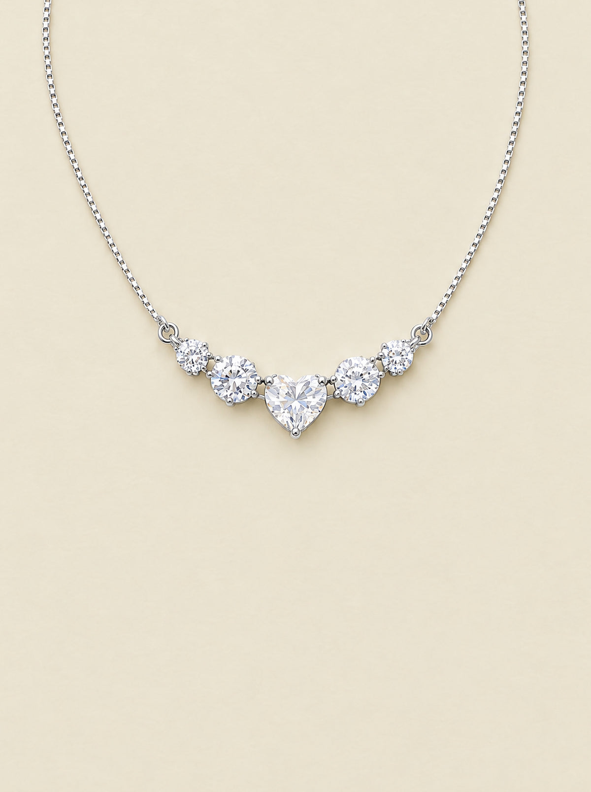 The Mossante Heart Necklace laid on a warm cream ground: a fine box chain in rhodium-white sterling silver, a short curved bar of five prong-set moissanites at the bottom, the heart at the center with two round brilliants stepping down on each side, the lobster clasp, extension chain and tiny heart tag at the top.