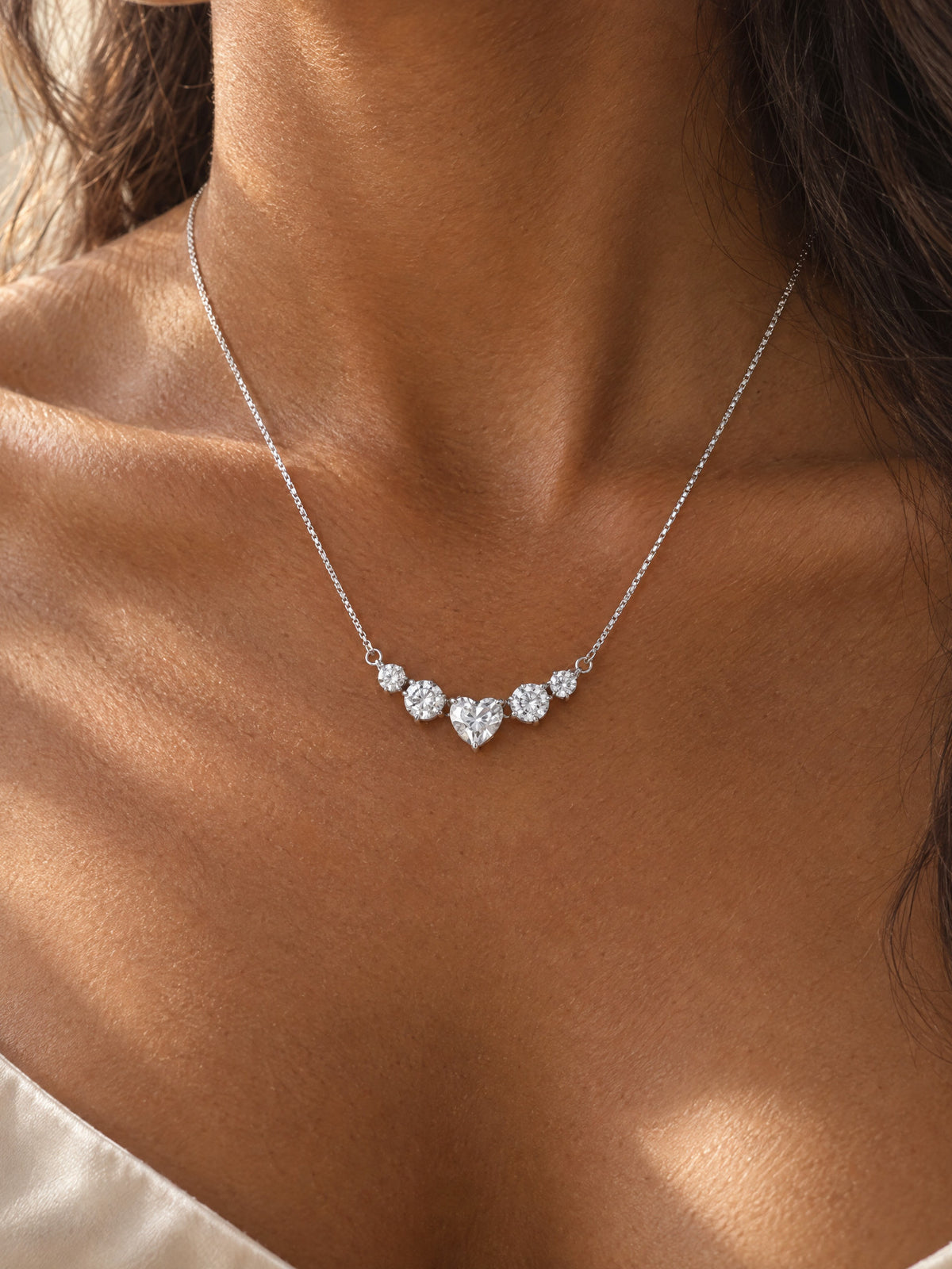 The silver heart necklace at the base of a woman's neck in warm window light, the heart and four rounds in one small curved row between the collarbones, a satin camisole strap at the shoulder.