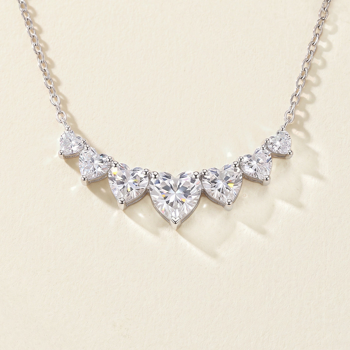 Close view of the seven heart-shaped moissanites in their curved row on a warm cream ground, each held in small silver claw prongs, the center heart the largest, light breaking into color at the facets.