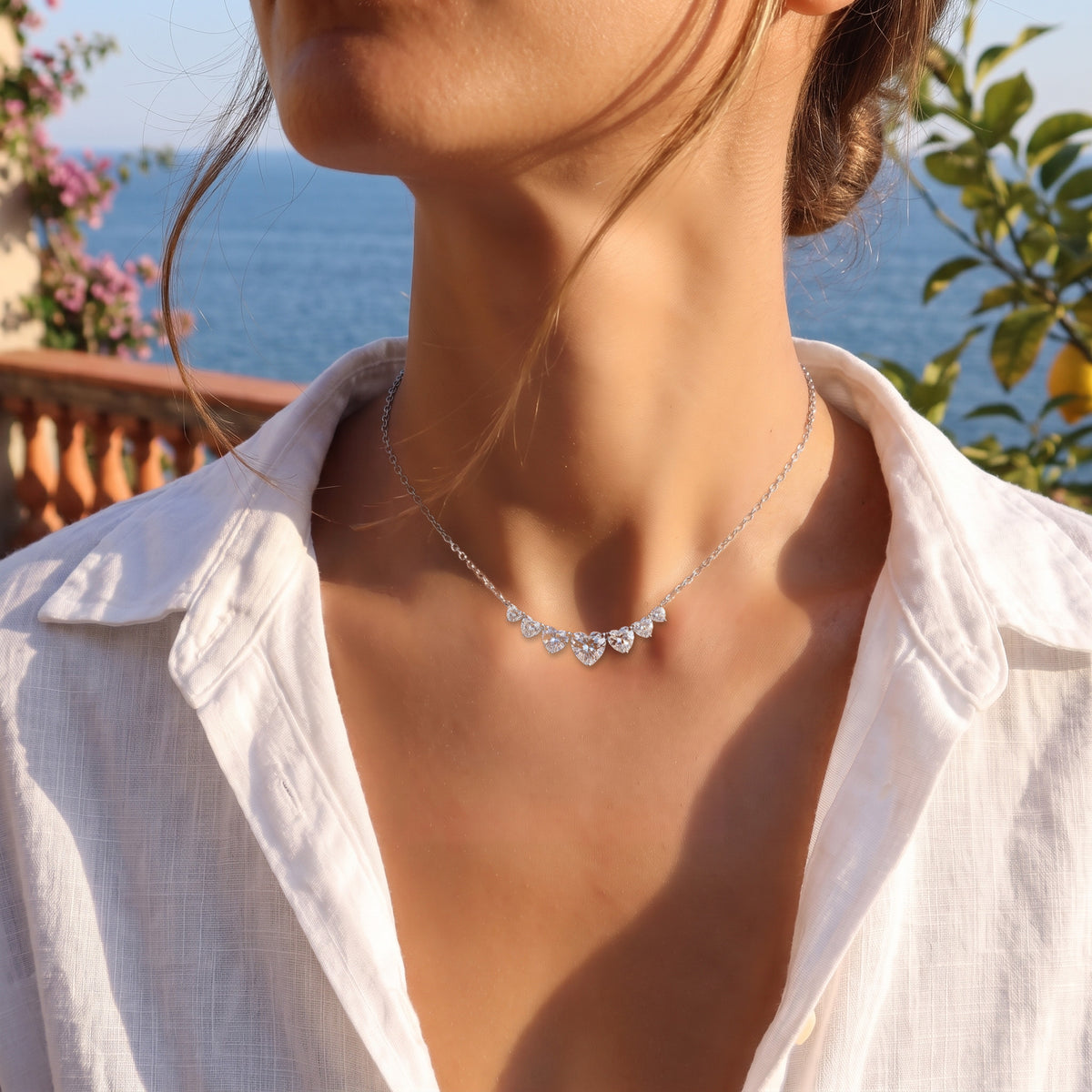 A sunlit terrace above the sea at golden hour, an open white linen shirt, wind in her hair, cropped at the chin.