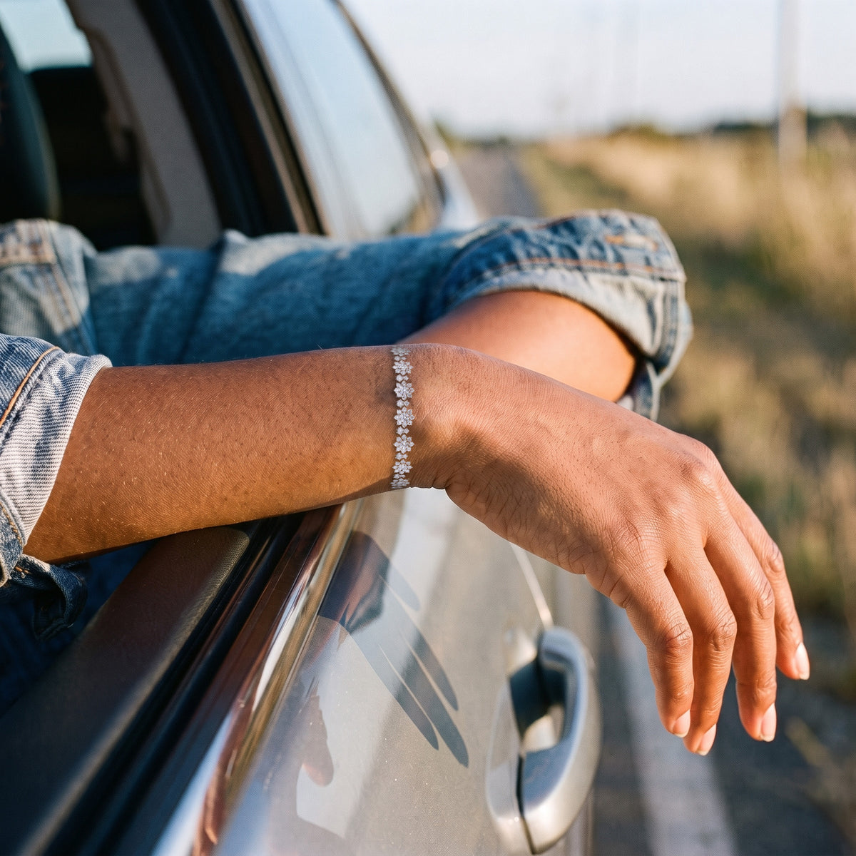 A woman's forearm resting on the open window of a car in late sun, a denim sleeve pushed back, the silver Bloom tennis bracelet lying across the top of her wrist, its flowers catching the light.