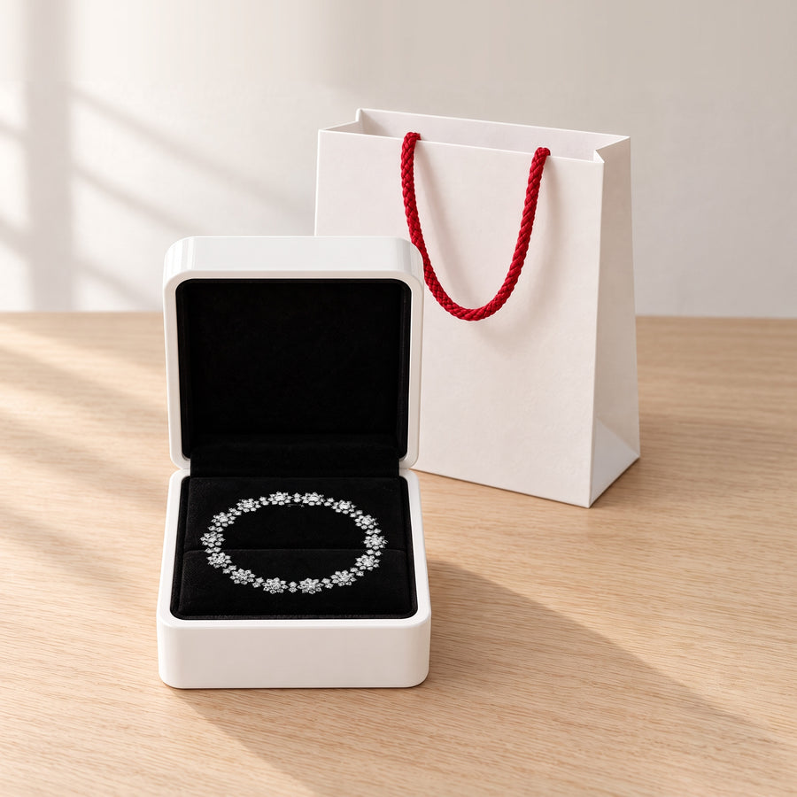 The silver Bloom tennis bracelet on the black velvet of its open white box, a white paper bag with red rope handles behind it on a pale wood table in window light.