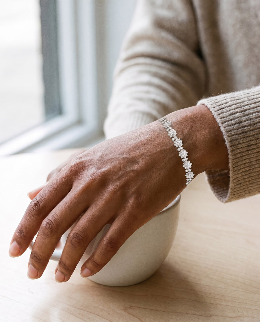 A woman with deep brown skin holding a plain white cup on a pale wood cafe table in morning window light, an oat knit sleeve pushed back, the silver Bloom tennis bracelet across the top of her wrist.