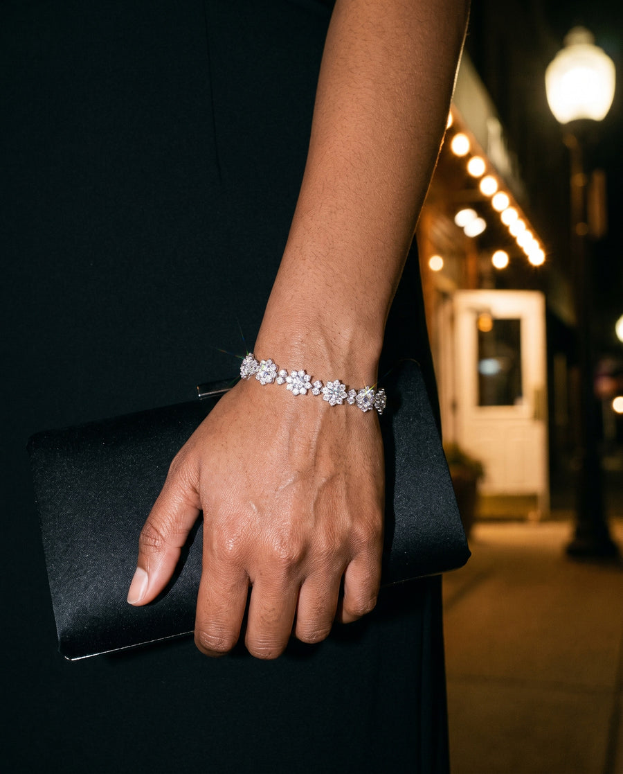 A South Asian woman's hand holding a black satin clutch against a black dress at a restaurant entrance at night, warm lights blurred behind, the bracelet's flowers catching the light at her wrist.
