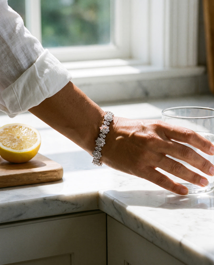 A woman with olive skin, a white linen cuff rolled back, her fingers round a glass of water on a white marble counter beside a halved lemon, bright daylight, the bracelet lying flat across her wrist.