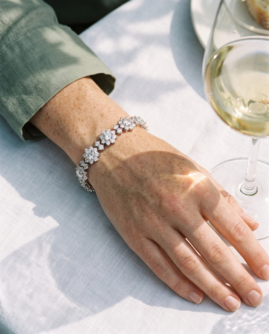 A light, freckled woman's hand resting on a white linen tablecloth beside a glass of white wine at a garden lunch in late sun, a sage linen sleeve, the flowers of the bracelet bright on her wrist.