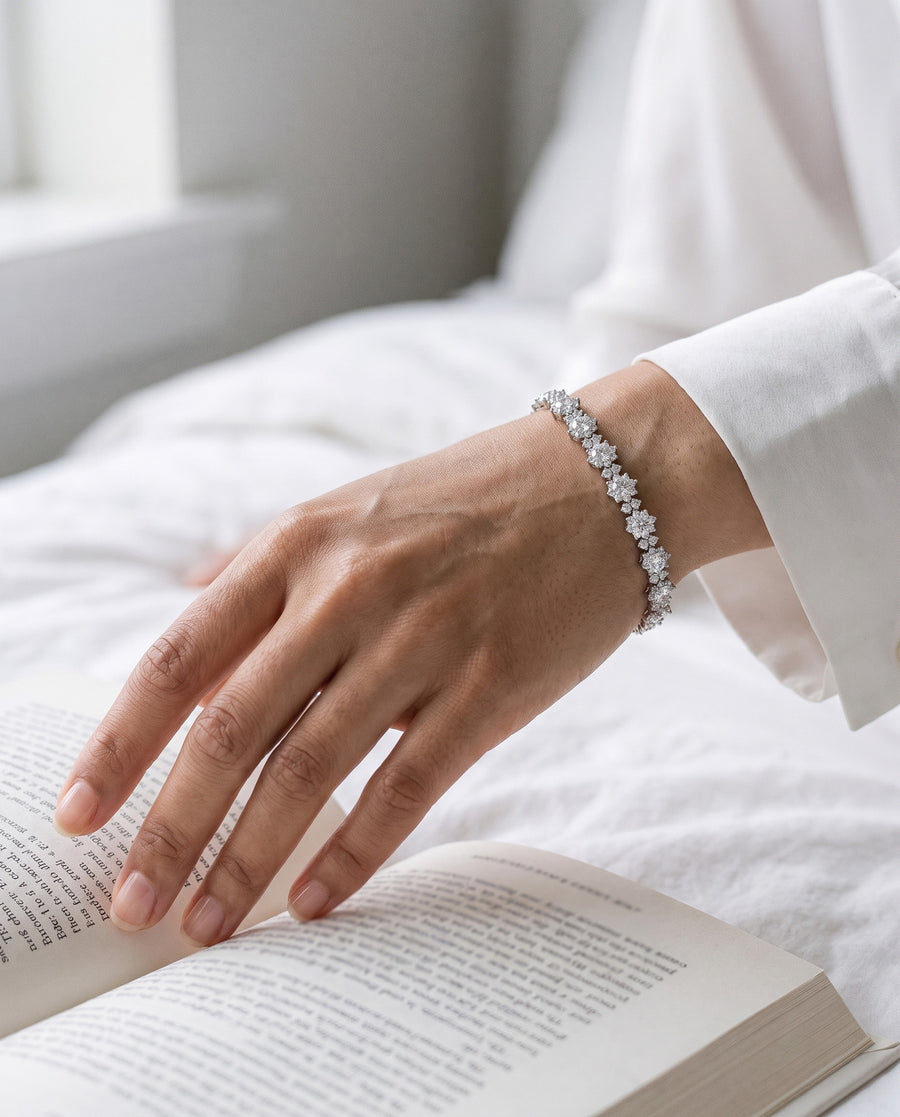 An East Asian woman's hand resting on the open page of a book on white bed linen in soft morning light, a white shirt cuff at her wrist, the silver tennis bracelet lying across it.