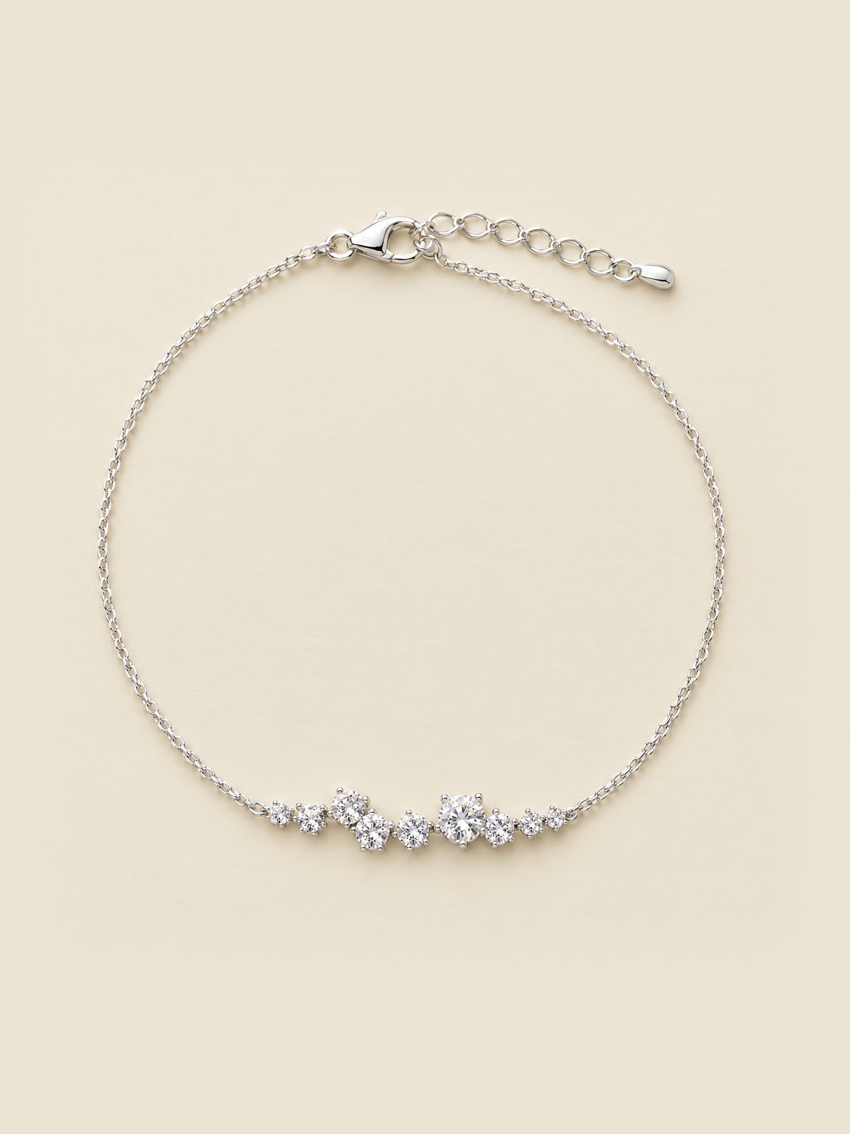 The Mossante Constellation Bracelet laid on a warm cream ground: a fine sterling silver cable chain with one small cluster of nine round brilliant moissanites at its center, the lobster clasp and extension chain to one side.
