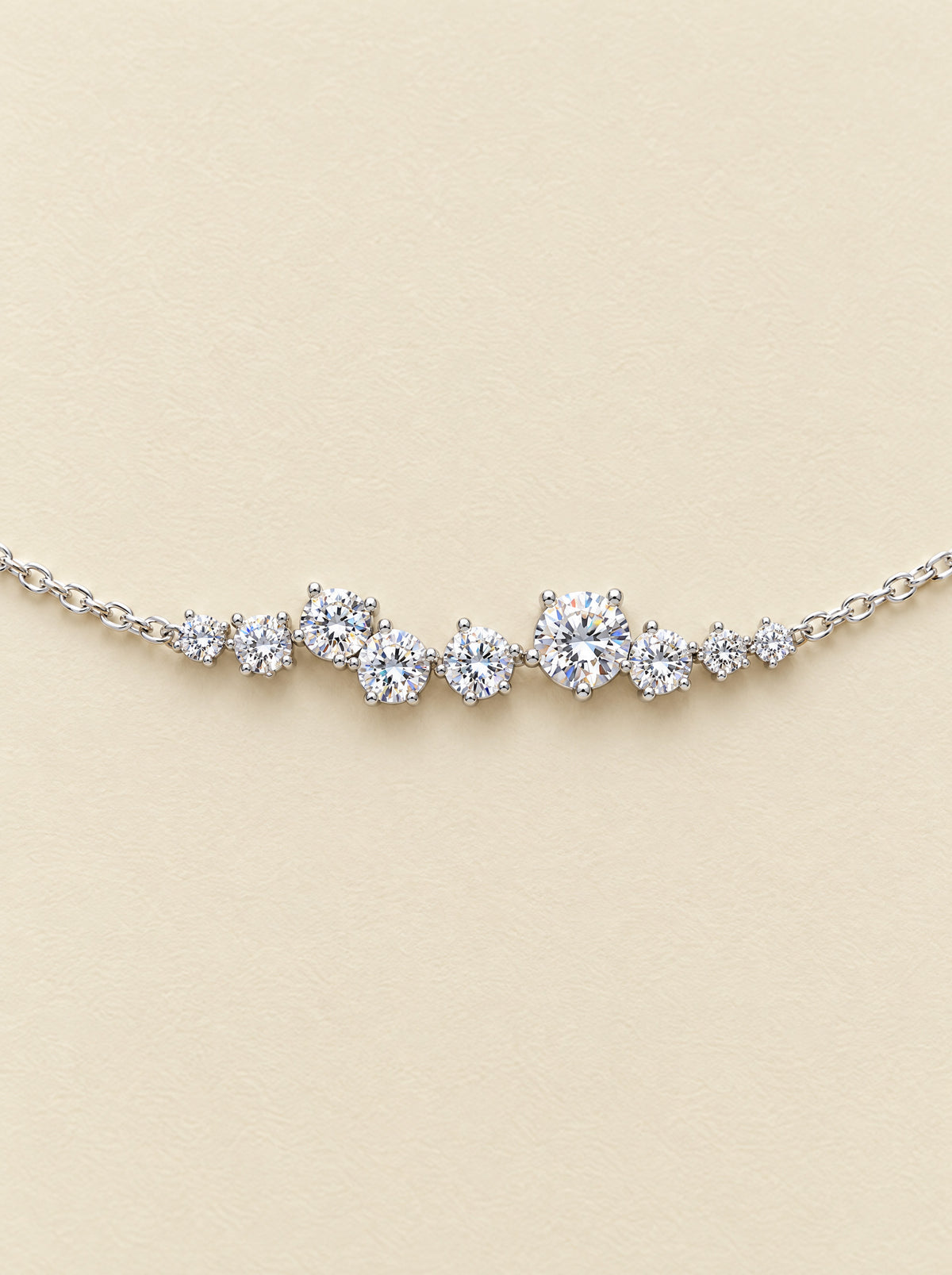 Close view of the cluster from straight above: nine round brilliant moissanites in three sizes, each in tiny prongs, lying along the fine sterling chain on a cream ground.