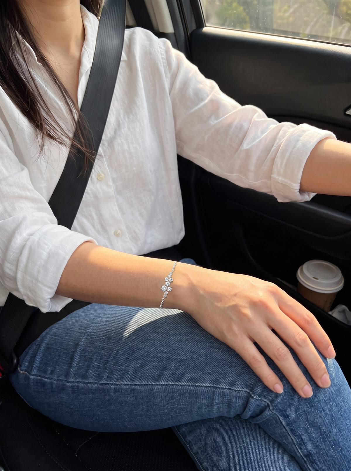 A woman in a white linen shirt in the passenger seat of a car, her hand resting on her knee, the nine-stone cluster on top of her wrist in warm window light.