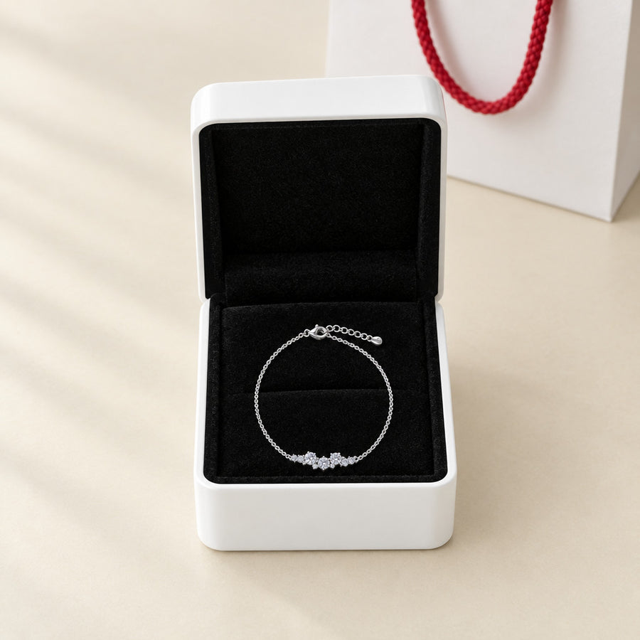 The constellation bracelet in its open white box with a black velvet interior, the white paper bag with red rope handles beside it, on a cream ground.