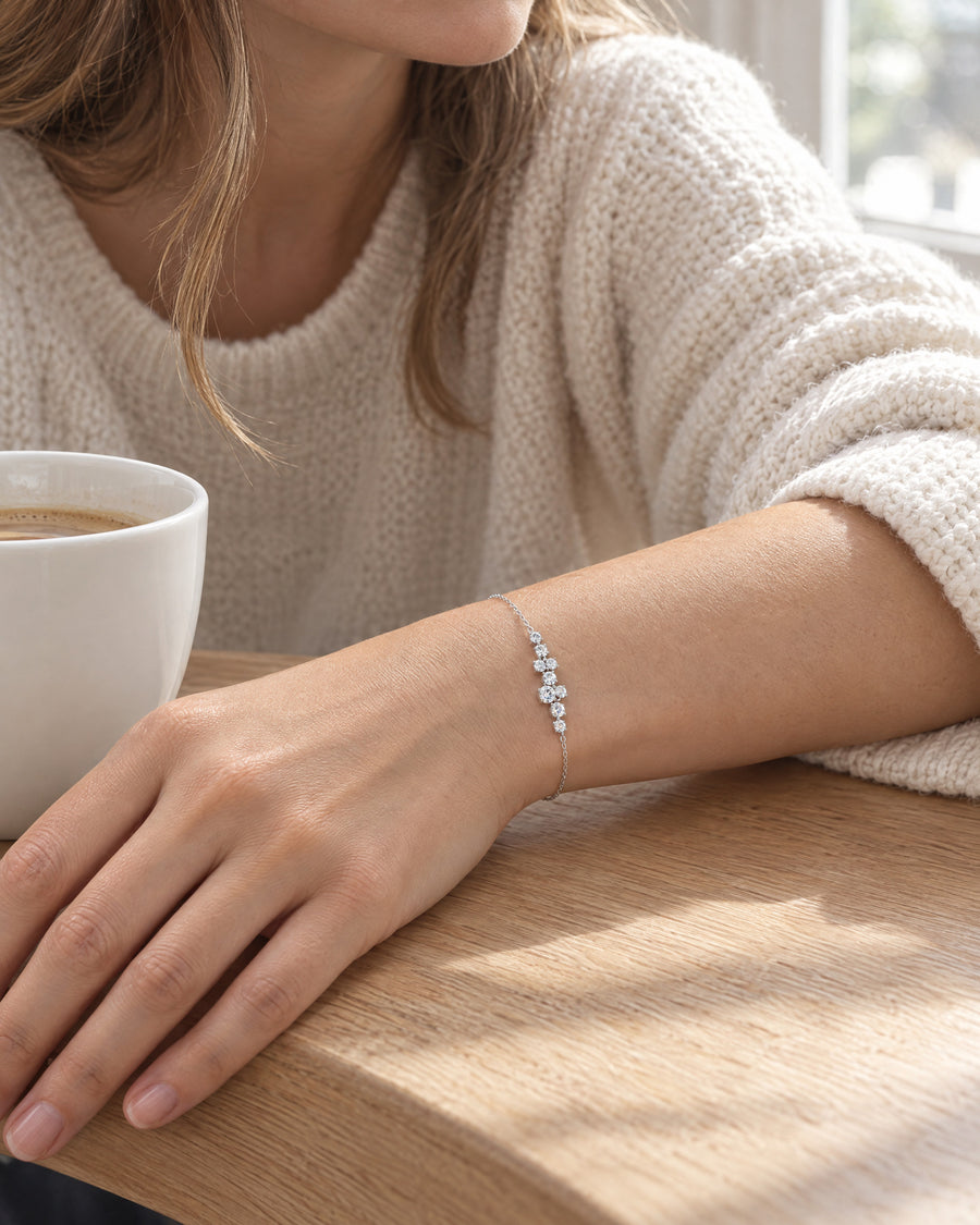 A woman's wrist resting on a light wooden cafe table beside a white ceramic cup in morning light, the constellation bracelet at the top of the wrist, a cream knit sleeve pushed back.