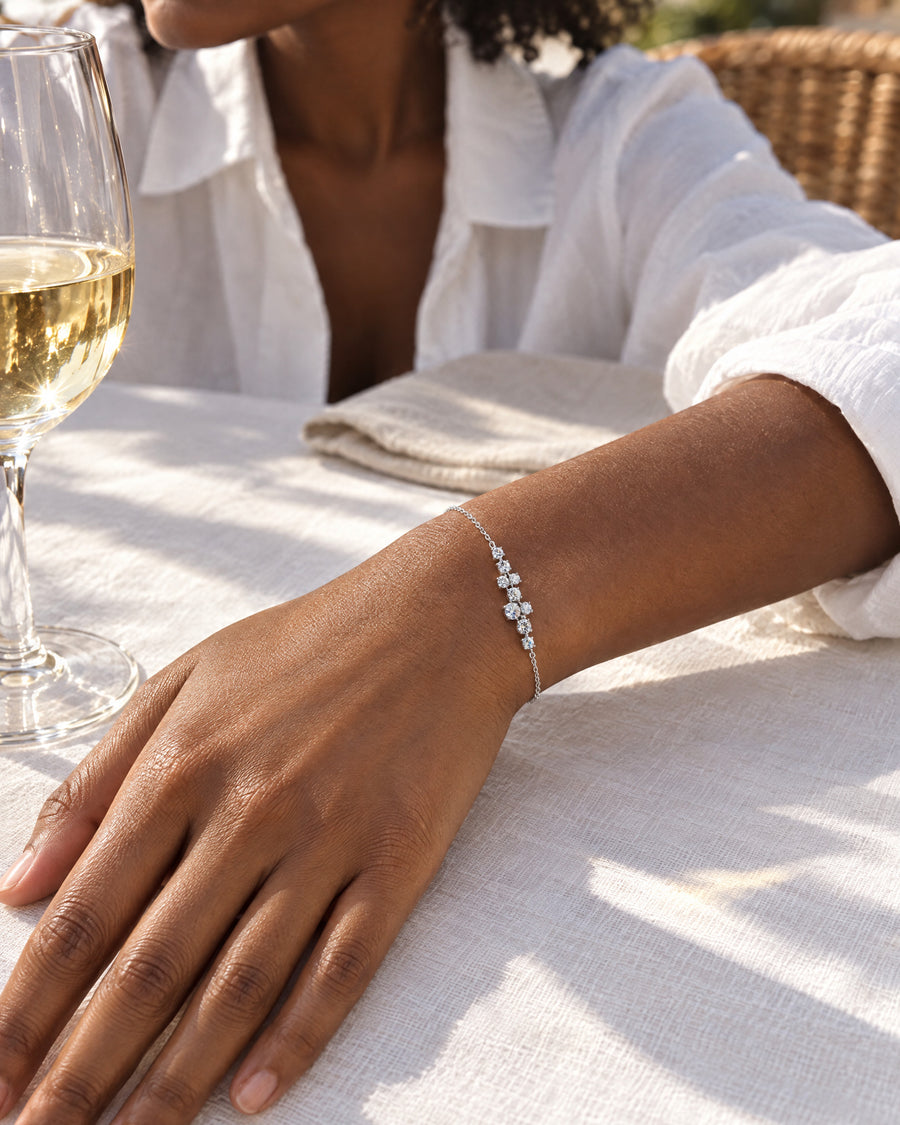 A Black woman's hand resting on a white linen tablecloth beside a glass of white wine in late afternoon sun, the bracelet loose on the wrist, the cluster catching the light.