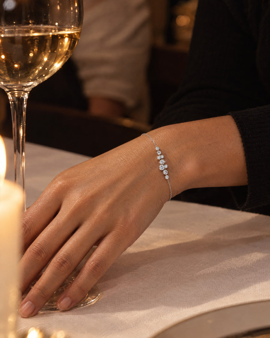 A woman's wrist on a white tablecloth beside the stem of a wine glass in candlelight, a black sleeve just visible, the nine stones the brightest points in the frame.