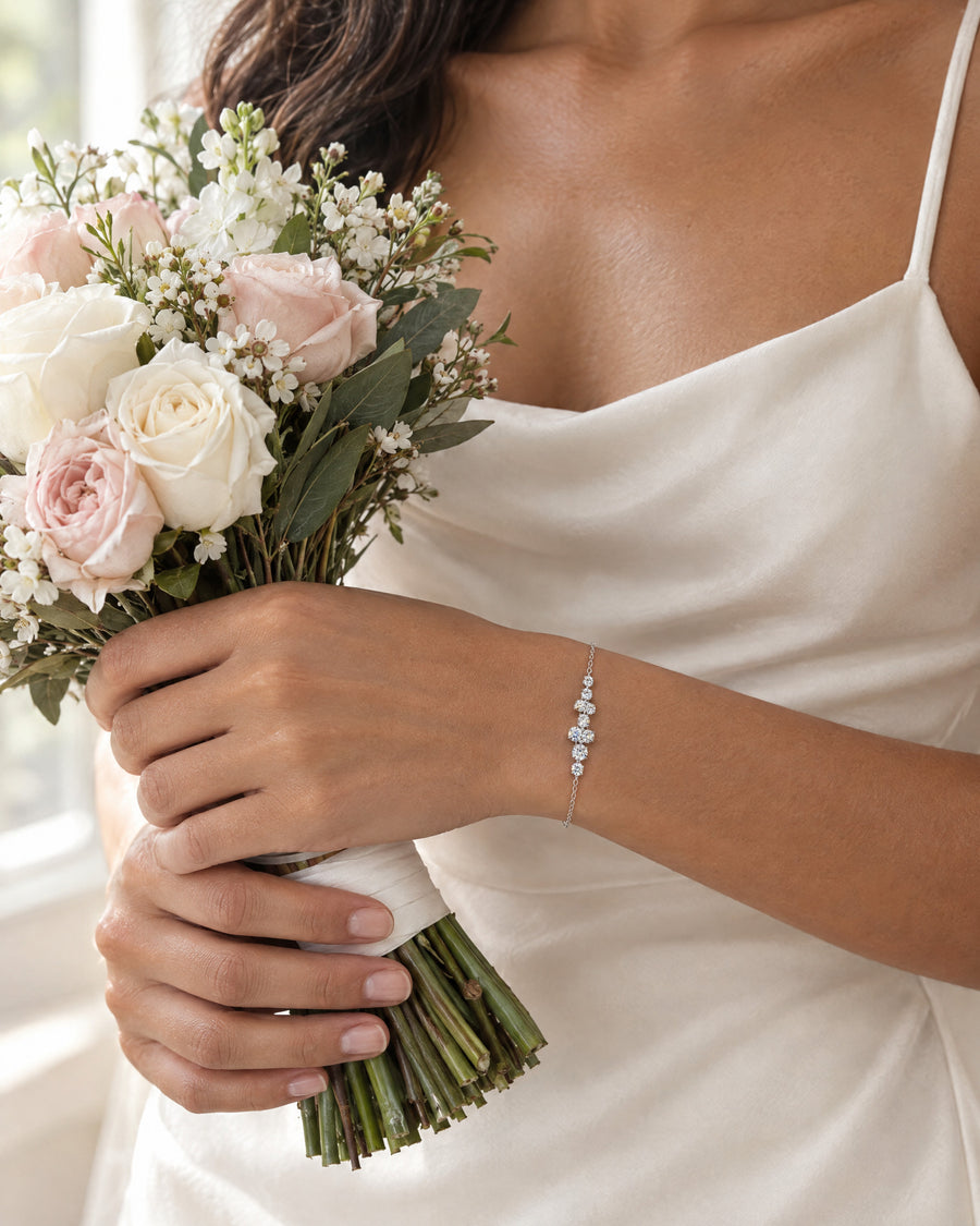 A bride's hand holding the stems of a small white bouquet against ivory silk, the constellation bracelet at the wrist in soft daylight.