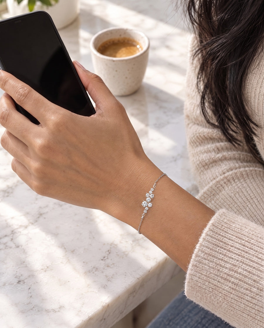 A South Asian woman's hand holding a black phone over a marble counter, an espresso behind, the bracelet lying at the wrist with the cluster facing up.