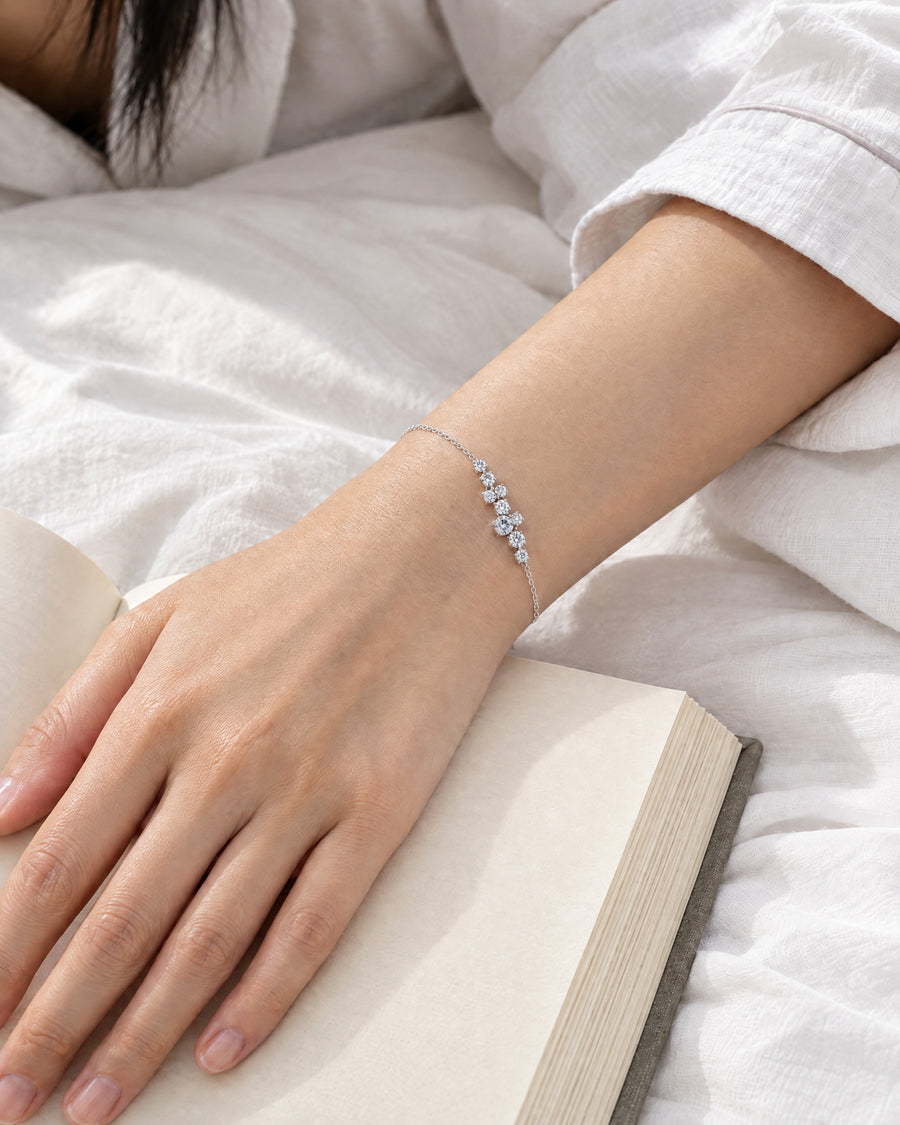 An East Asian woman's hand resting on the open page of a book on white bed linen in morning light, the nine-stone cluster on top of the wrist, the fine chain running round it.
