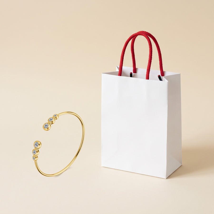 The gold plated Bezel Cuff on a cream ground beside a white paper gift bag with red rope handles.