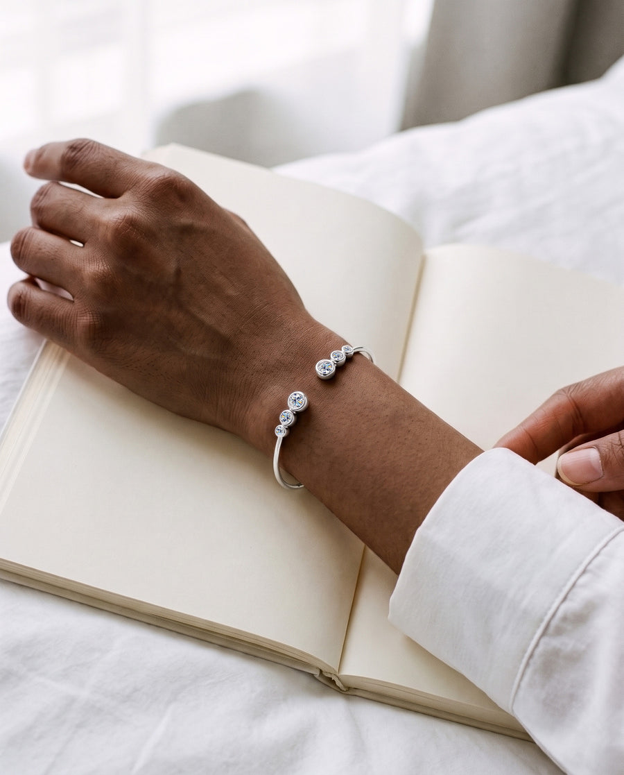 A woman's hand resting on an open notebook on white bed linen in soft morning light, a white shirt cuff beside it, the silver Bezel Cuff on top of her wrist.