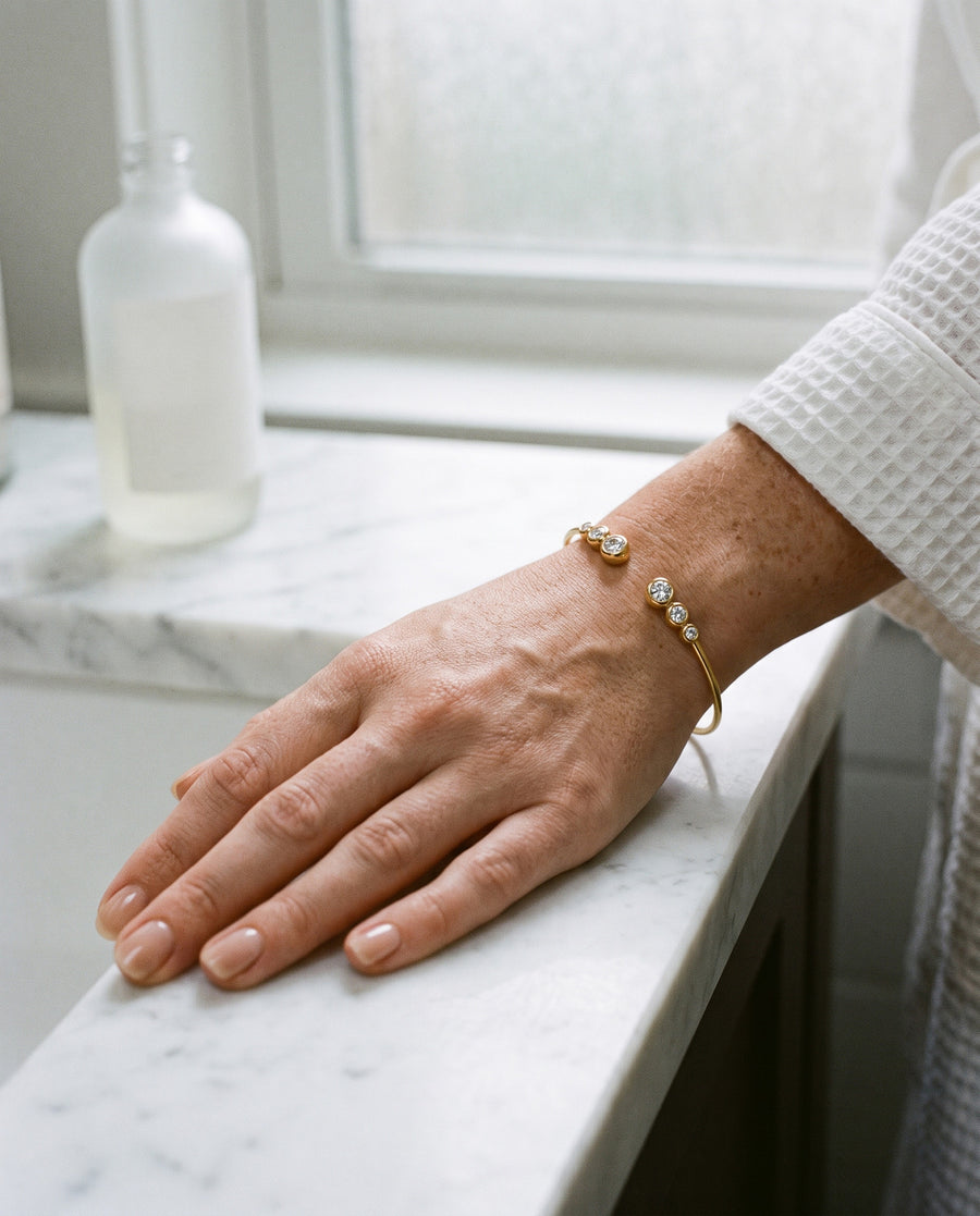 A light-skinned, freckled woman's hand resting on a white marble bathroom counter beside a frosted unlabeled bottle, a white waffle robe sleeve at the wrist, the gold plated cuff's six stones on top of her wrist.