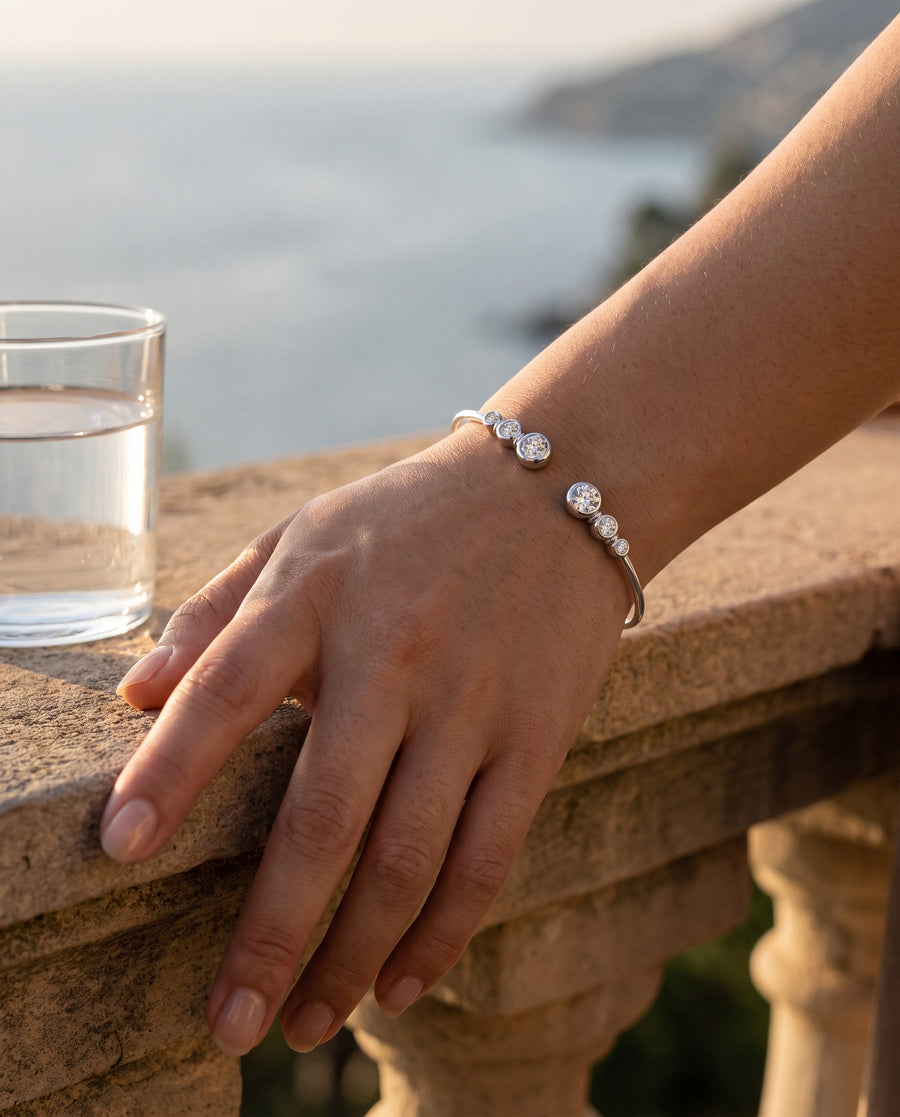 An East Asian woman's bare forearm resting on a warm stone balcony ledge in late sun beside a glass of water, a soft sea haze behind, the two stone ends of the silver cuff catching the light on top of her wrist.
