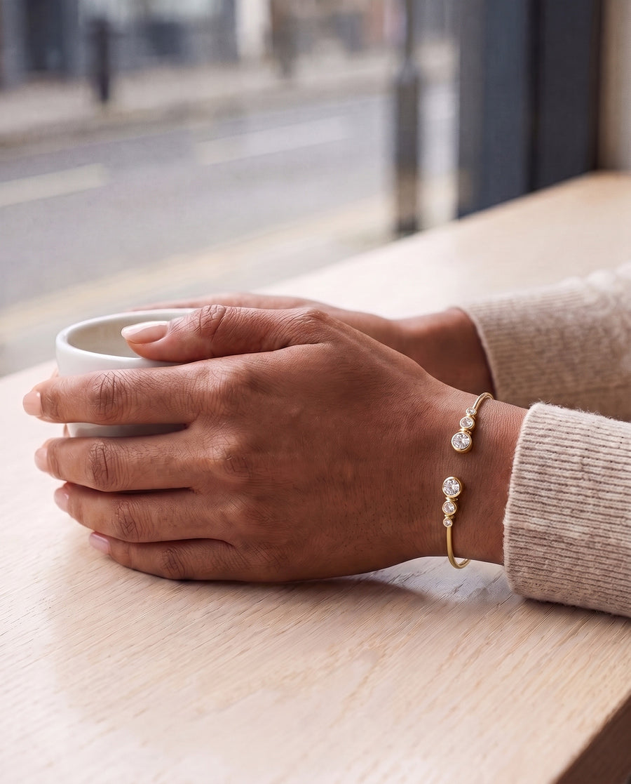 A woman's hands round a white cup at a cafe window, an oat knit sleeve pushed back, the gold plated Bezel Cuff on top of her wrist.