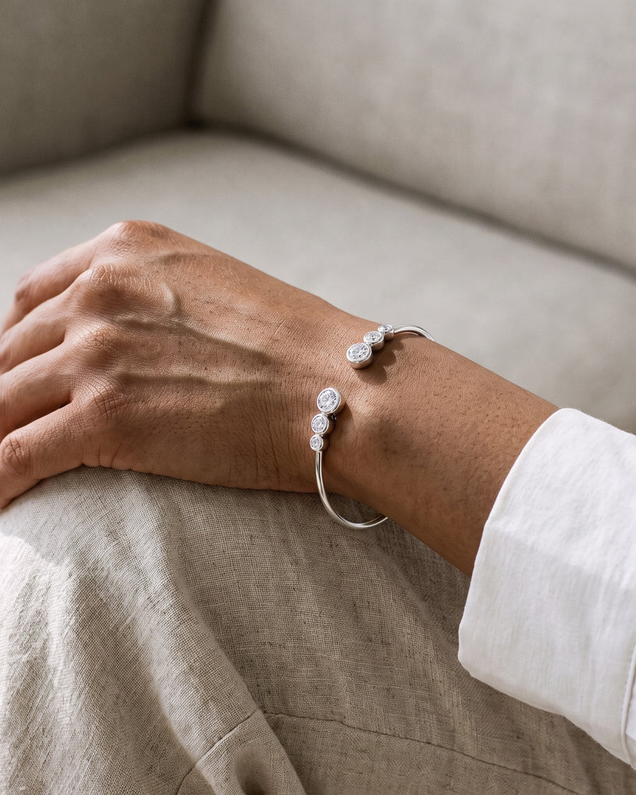 A woman's hand resting on her knee on a linen sofa, a white shirt cuff rolled back, the silver Bezel Cuff's two stone ends on top of her wrist.