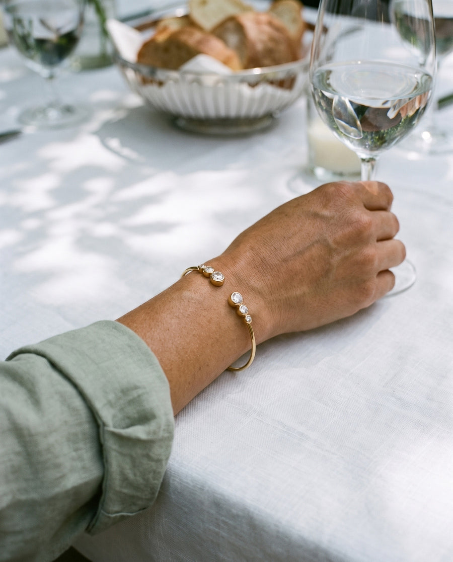 A woman's hand holding a glass of water at a garden lunch on white linen in dappled shade, a sage sleeve, the gold plated Bezel Cuff at her wrist.
