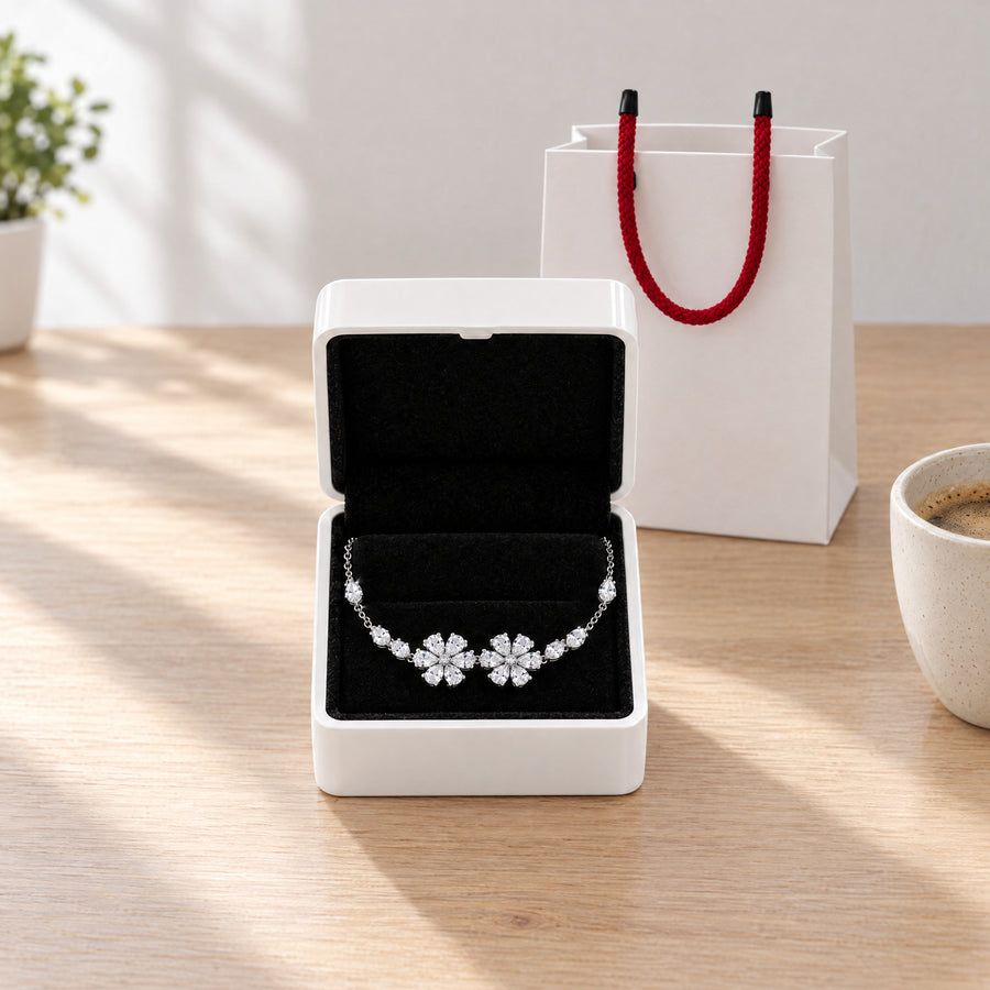 The floral bracelet in its white box with the lid open, lying on black velvet, the white gift bag with red rope handles behind.