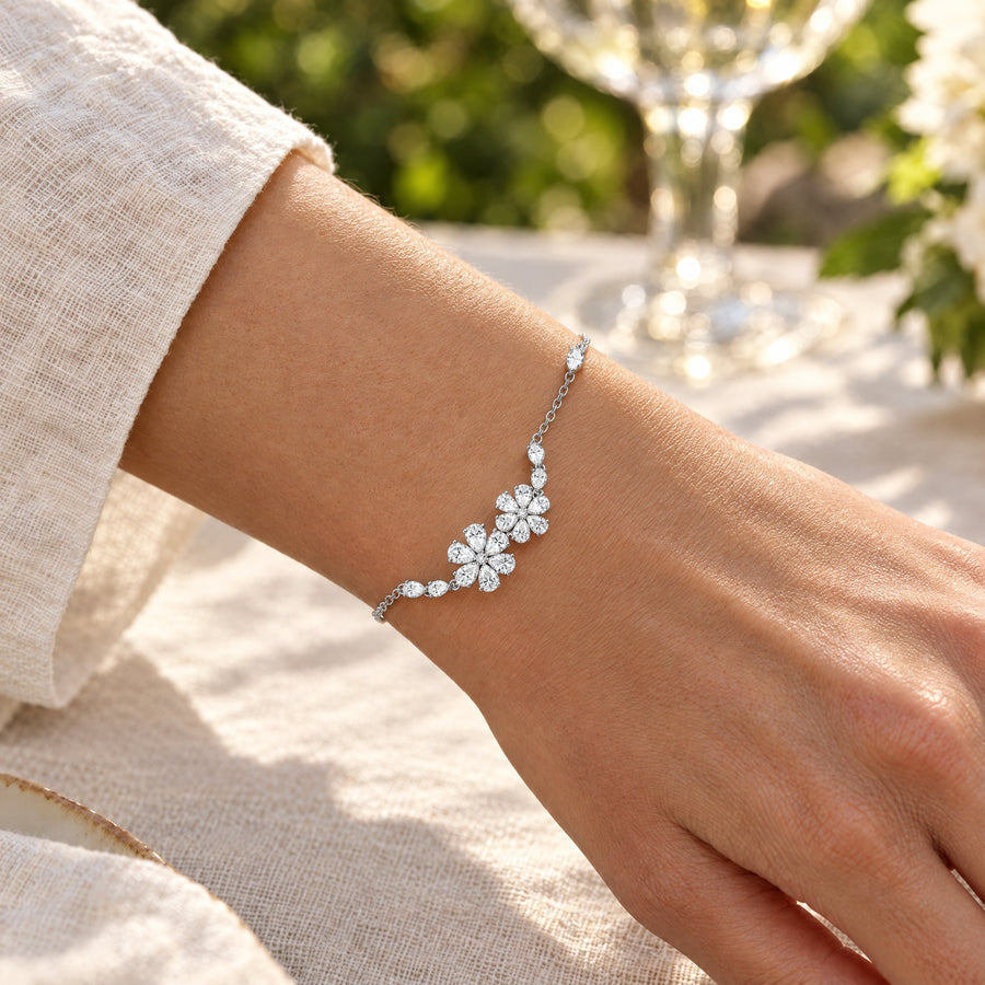 A woman's wrist at an outdoor brunch table beside a rolled cream linen sleeve, the floral bracelet catching the sun, a glass and white flowers out of focus beyond.