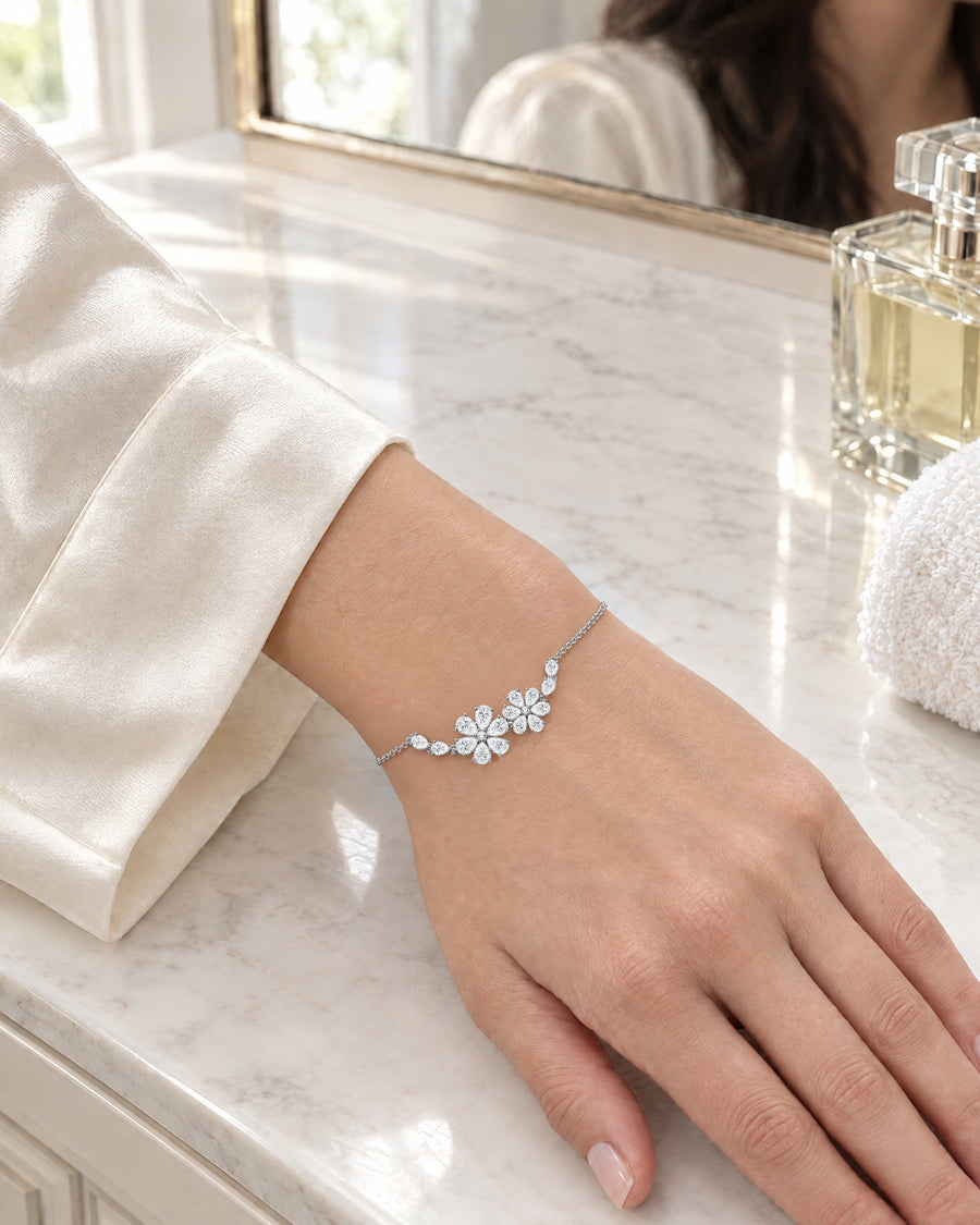 A woman's hand resting on a white marble bathroom counter in morning light, an ivory silk robe sleeve at the wrist, the two flowers facing up beside an unlabelled perfume bottle.