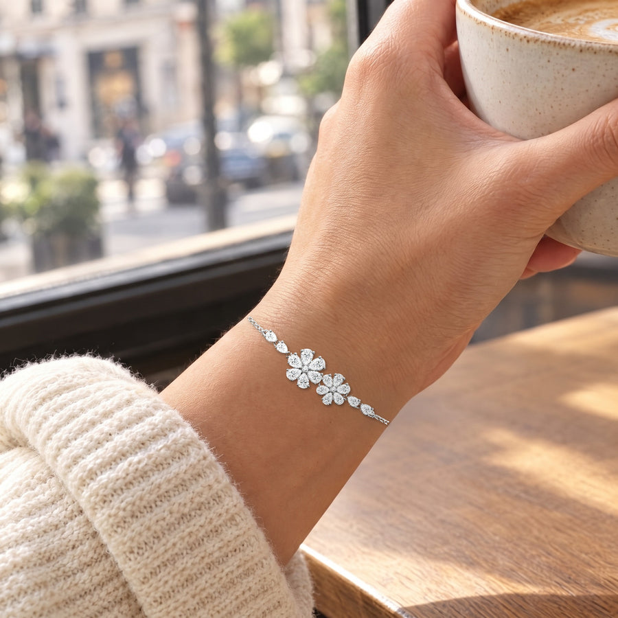 A woman's hand around a café cup by a window, a cream knit sleeve at the forearm, the floral bracelet on the wrist and the flowers turned to the light.