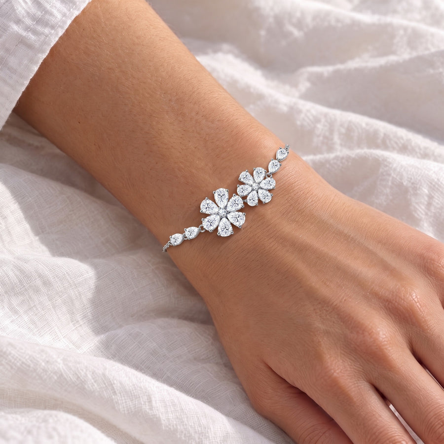A woman's forearm and hand resting on white hotel bed linen in morning light, the floral bracelet on the wrist with both flowers facing up.