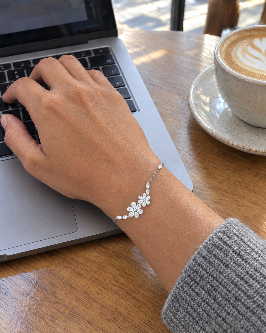A hand typing on a laptop at a café table, the floral bracelet on the wrist and a latte beside it.
