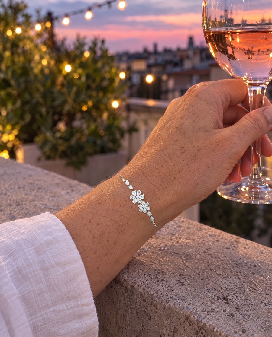 A hand holding a glass of rosé on a rooftop at dusk, string lights behind, the floral bracelet on the wrist.