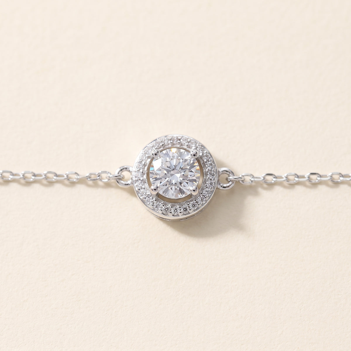 Close view of the halo from above: one round moissanite at the center, ringed by tiny stones inside a round silver rim, the fine cable chain joining it at each side, on a cream ground.