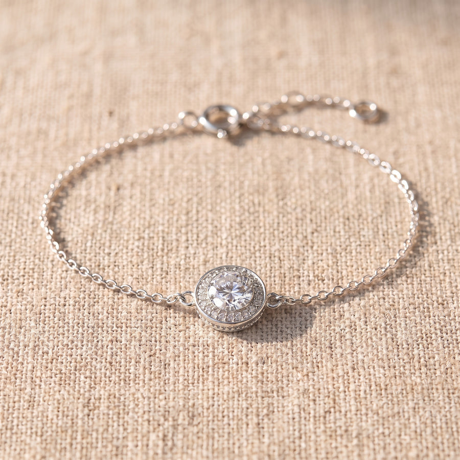 Close view of the halo from a low three-quarter angle on oat linen: the round center stone raised above a ring of tiny stones in a round silver rim, the fine cable chain curving away on both sides.