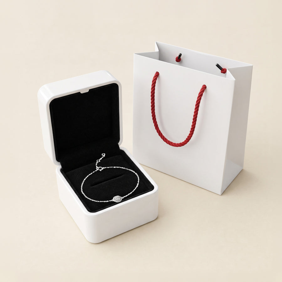 The Halo bracelet in its open white box, the halo resting on the black velvet interior, the white paper bag with red rope handles beside it on a cream ground.