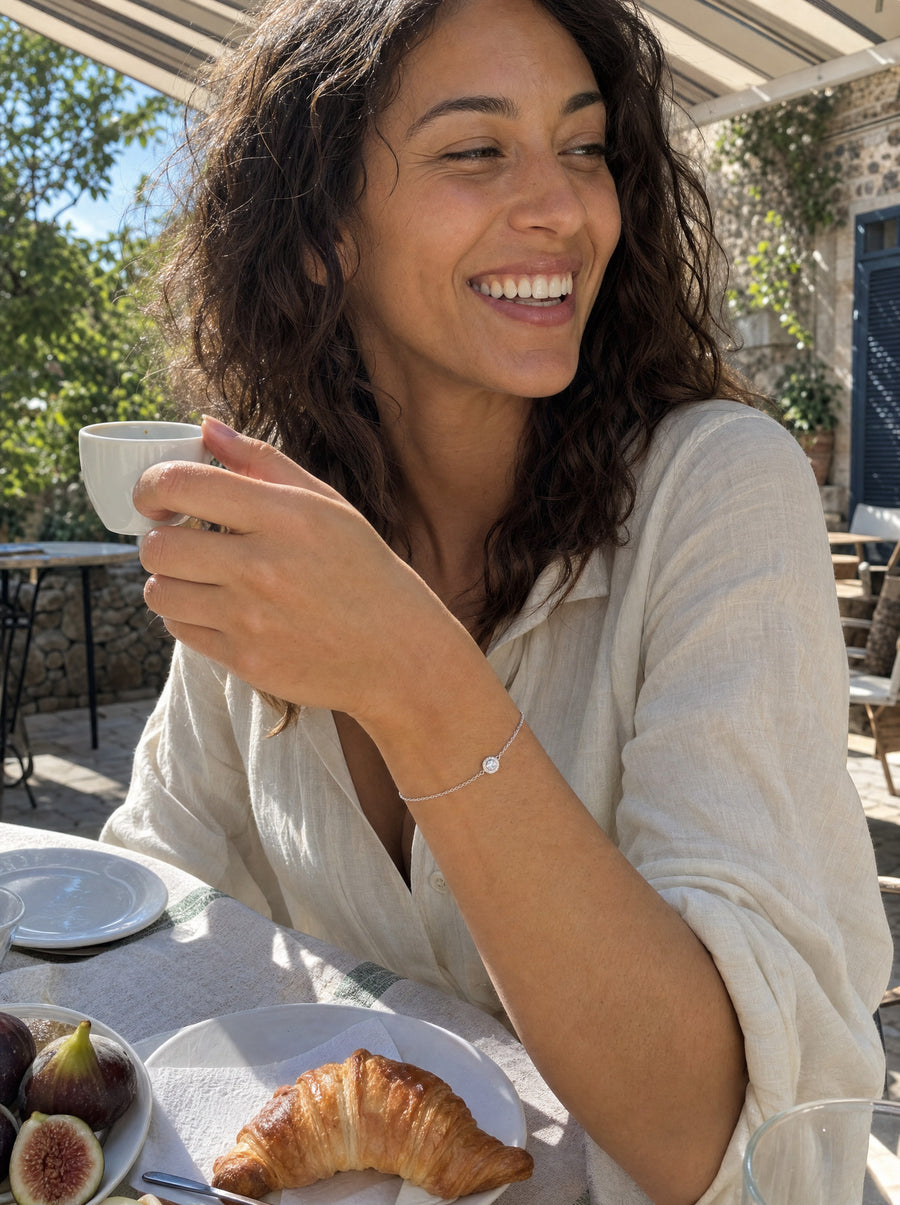 A young woman laughing at a sunny cafe table under a striped awning, lifting an espresso cup, the halo bracelet at her wrist.