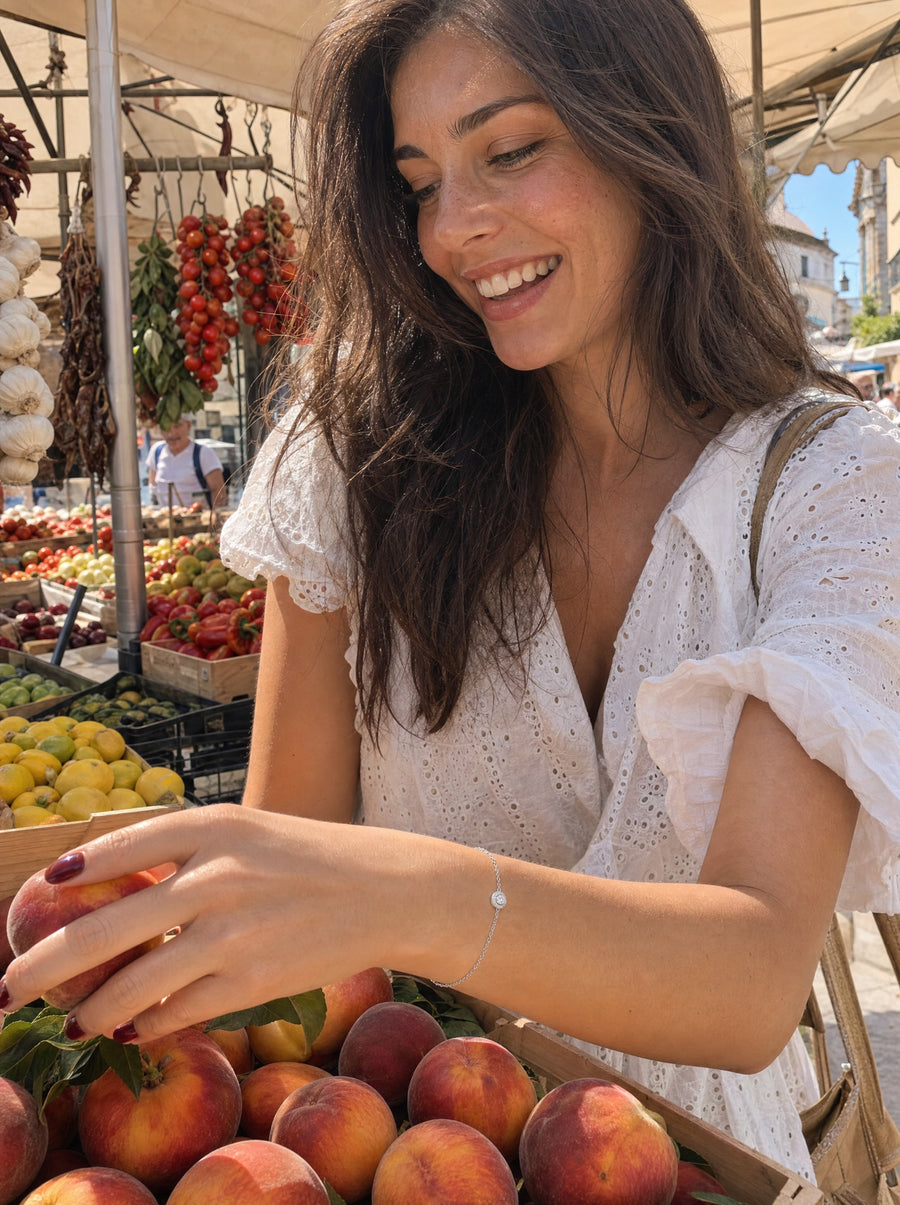 A young woman smiling as she chooses ripe peaches at a summer market, the halo bracelet at her wrist.