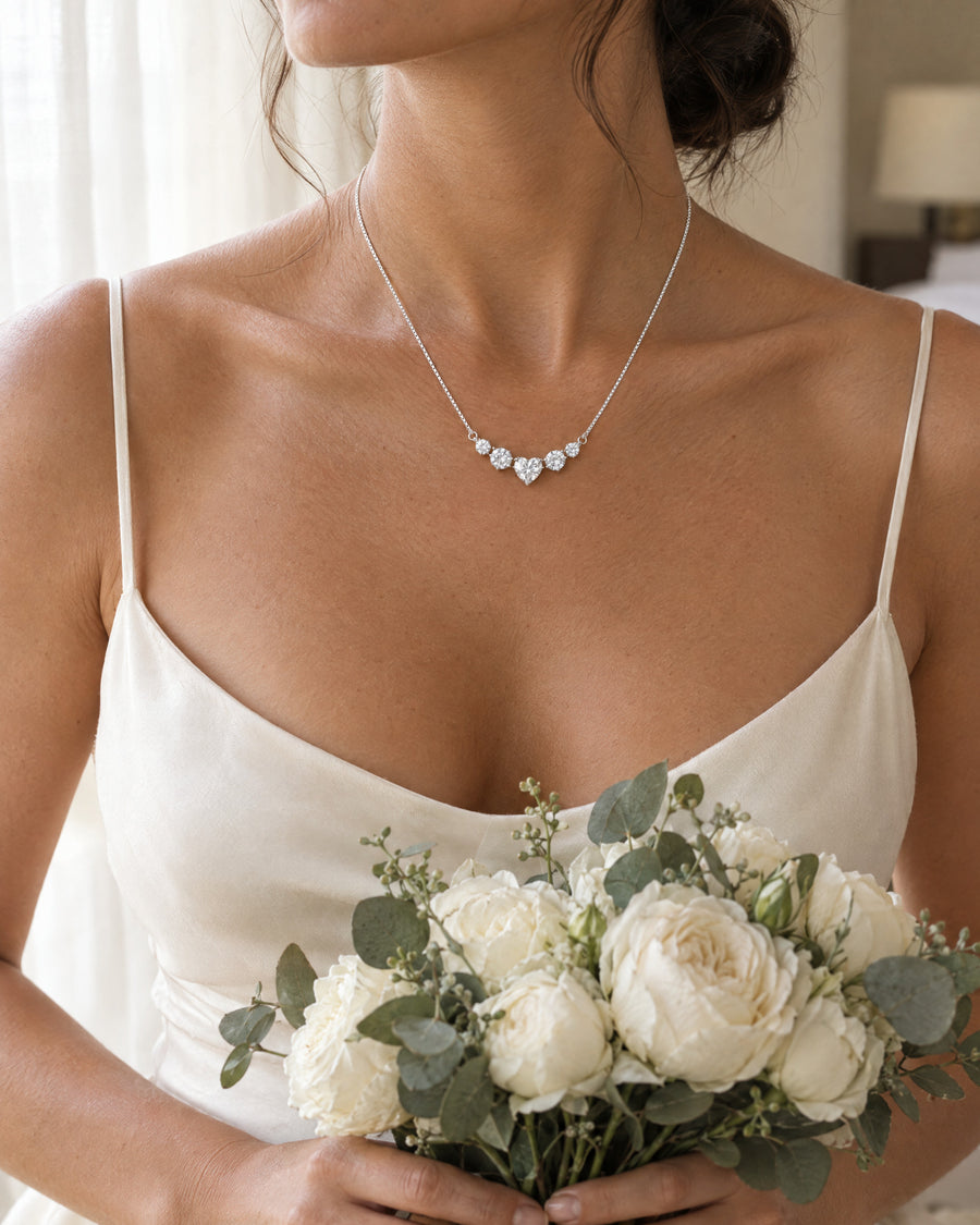 A bride's collarbone above an ivory silk neckline, the silver heart necklace lying in a gentle curve, the five stones catching daylight, a white bouquet held below.