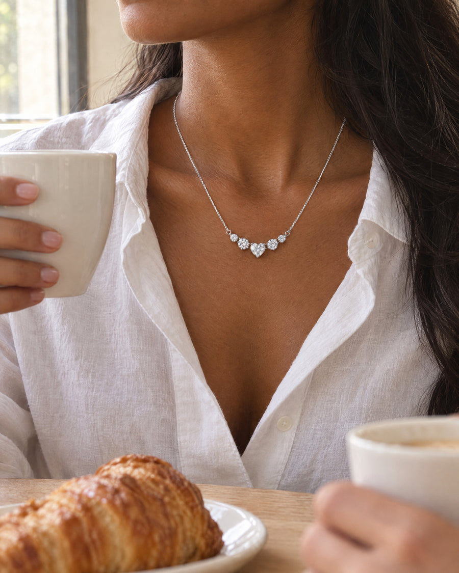 A woman at a light wooden cafe table in morning light, a white shirt open at the collar, the silver necklace resting at her collarbone, a white cup in her hand, cropped at the chin.