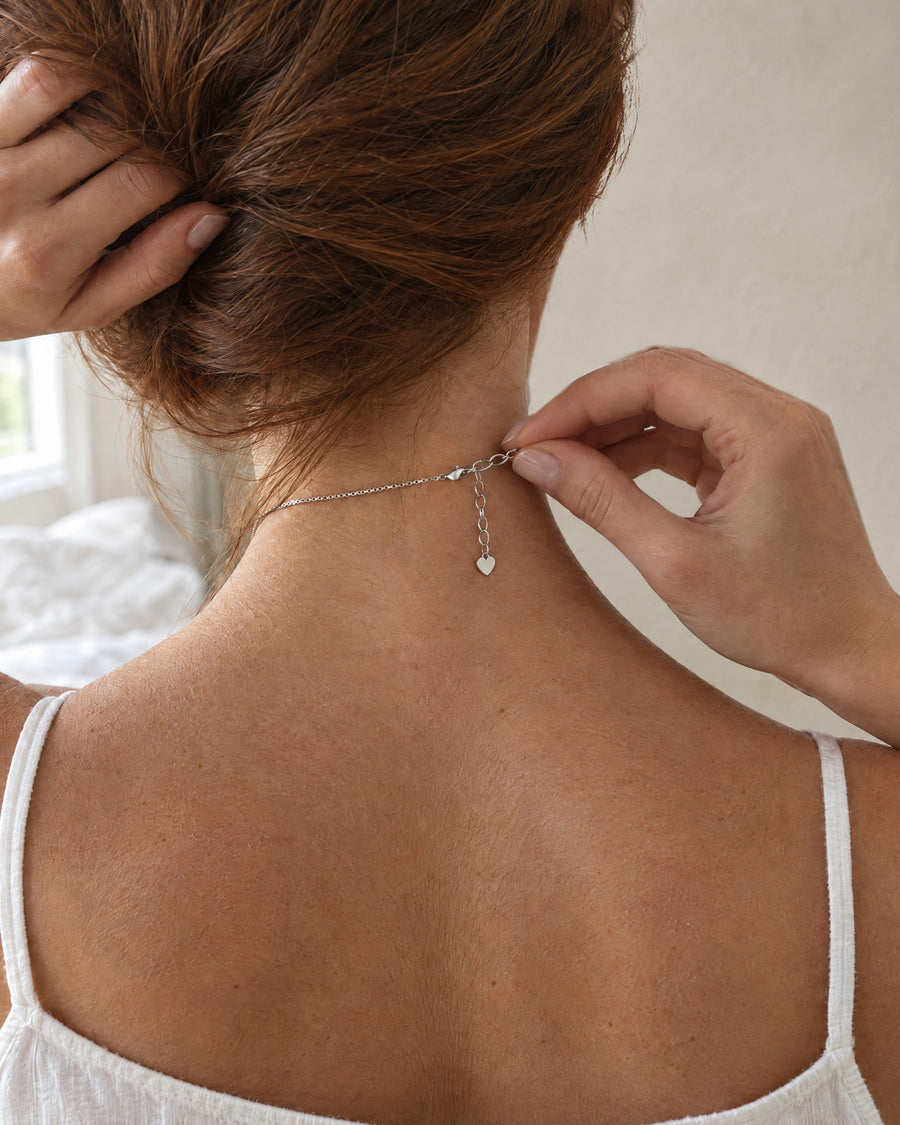 A woman seen from behind fastening the lobster clasp of the silver necklace at the back of her neck, hair pinned up, a plain limewashed wall beside her in soft morning light.