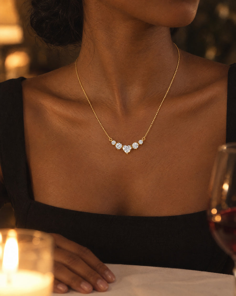 A woman in a black dress at a dinner table in candlelight, the gold-plated necklace at her collarbone, the heart the brightest point in the frame.