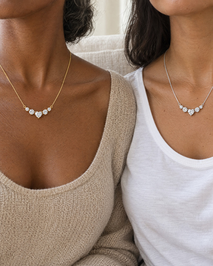 A mother and a daughter side by side on a linen sofa in daylight, one wearing the gold-plated necklace and one the silver, both cropped at the chin.