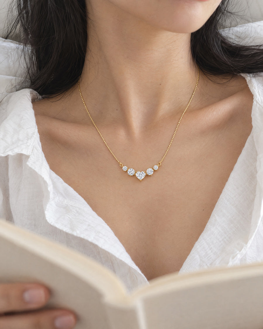 An East Asian woman in a loose white shirt resting against white bed linen with an open book, the gold-plated necklace loose at the collarbone, cropped at the chin.