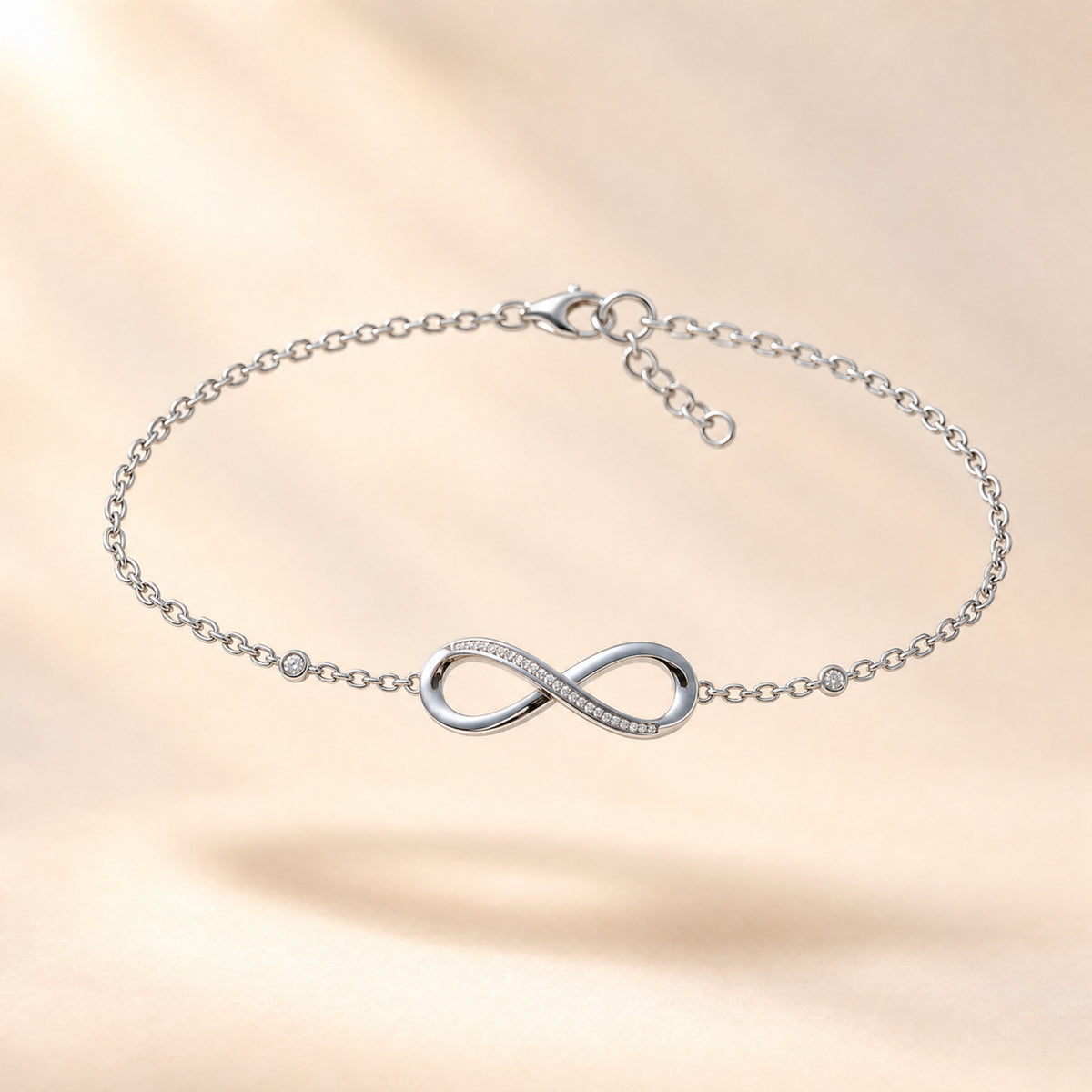 The Mossante Infinity Bracelet floating on a warm ivory ground: a fine silver chain, a polished infinity figure with a row of tiny stones, and a small round stone on the chain on each side.